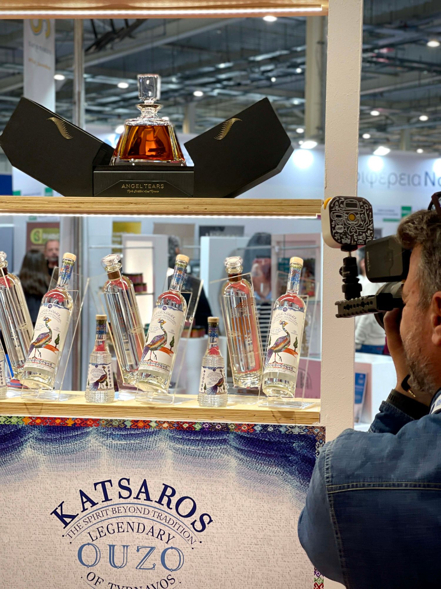 Το περίπτερο της Katsaros σε έκθεση με το Angel Tears και τα μπουκάλια ούζο, ενώ ένας κάμεραμαν κινηματογραφεί