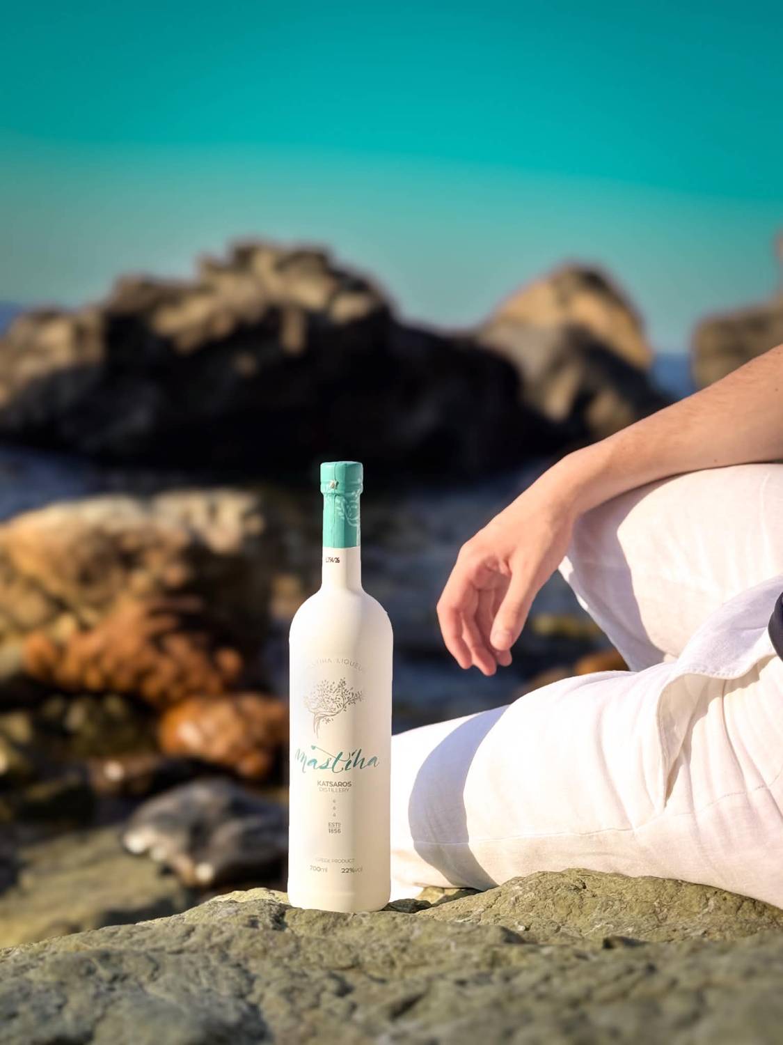 Λευκό μπουκάλι λικέρ μαστίχας Katsaros Distillery σε βράχο δίπλα στη θάλασσα, δίπλα σε άνθρωπο με λευκό λινό ρούχο