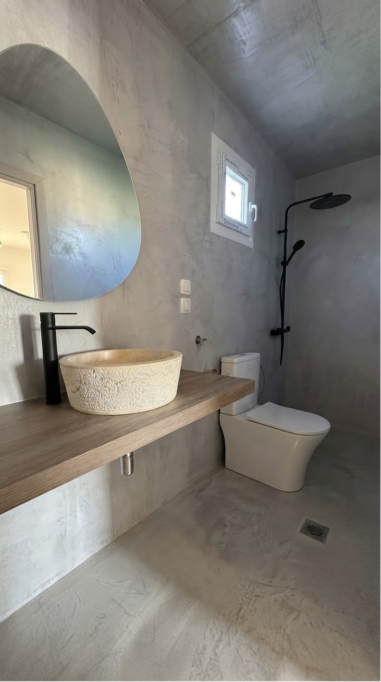 A bathroom with grey microcement, a black tap and shower, a stone basin on a wooden shelf and an oval mirror
