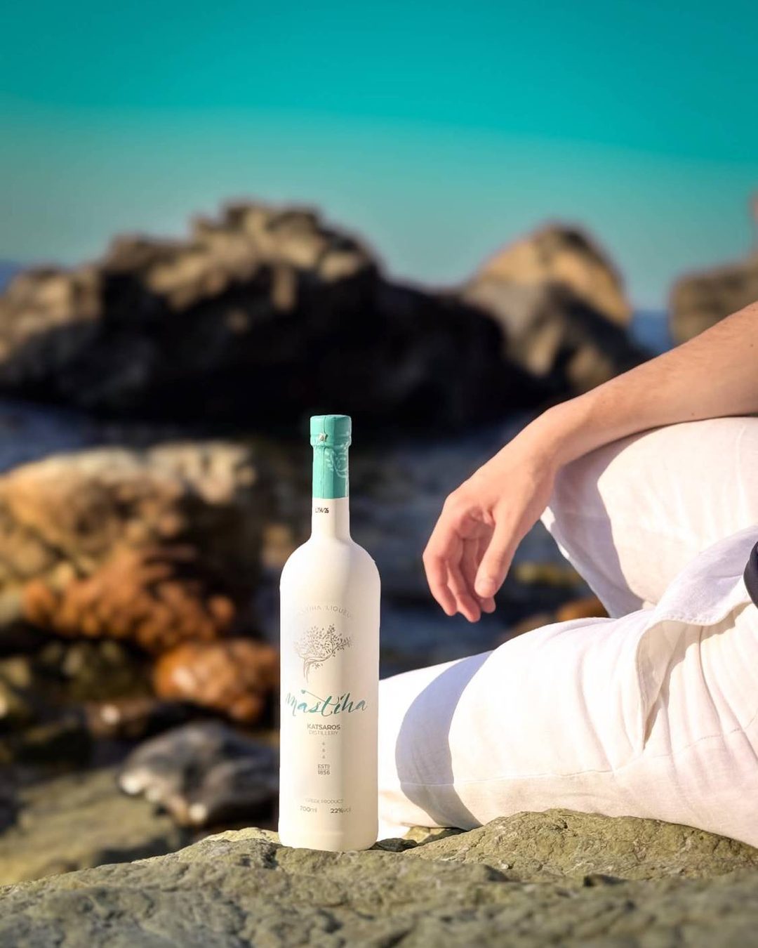 Katsaros Distillery Mastiha liqueur bottle on a seaside rock beside a person in white linen, with a turquoise sea behind